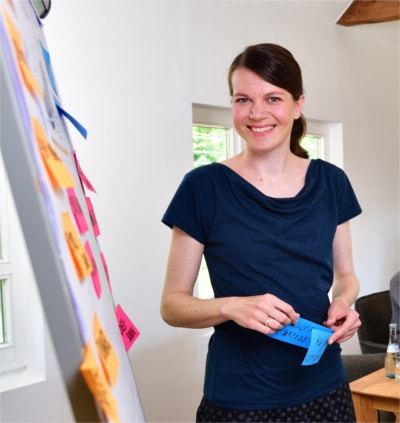 Eine Frau mit einem größeren Post-It steht vor einem Whiteboard.