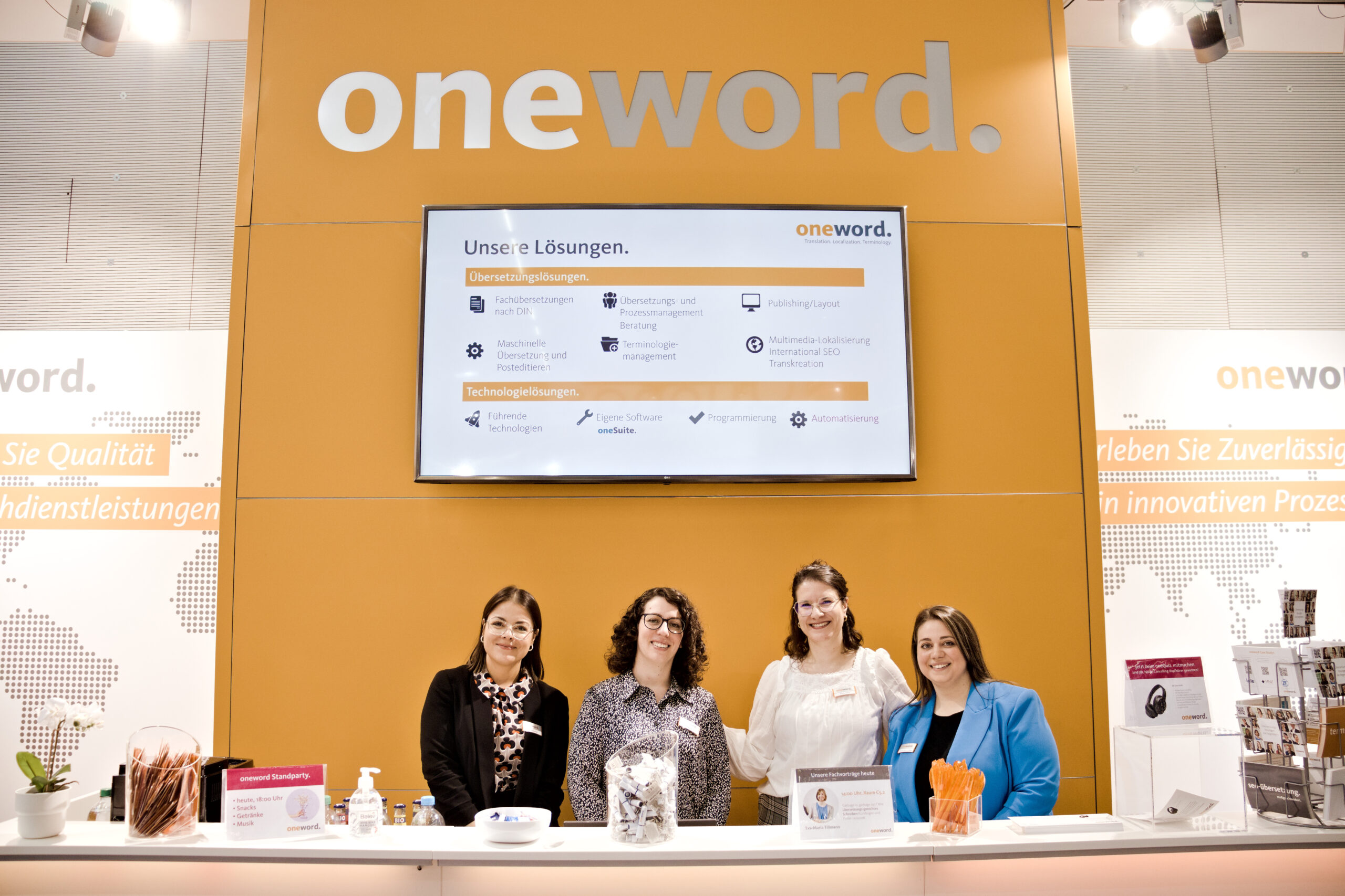 tekom Jahrestagung 2023 - oneword Messestand; 4 Frauen stehen unter einem großen Bildschirm und dem oneword Logo